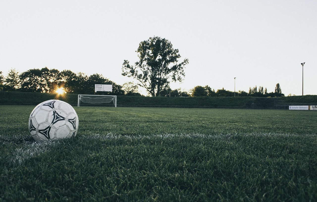 Ballon de football posé sur une pelouse sombre au coucher du soleil, un but en arrière-plan