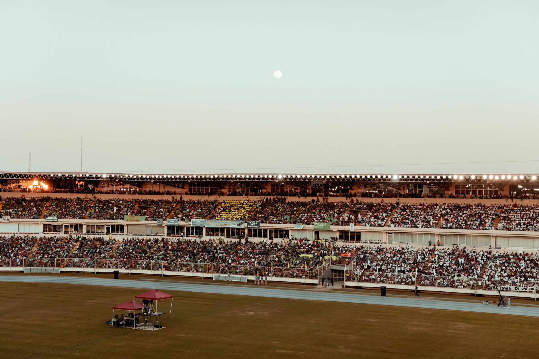 Stade au crépuscule, tribunes pleines de spectateurs sous des projecteurs allumés, piste bleue et pelouse au premier plan, lune dans le ciel.