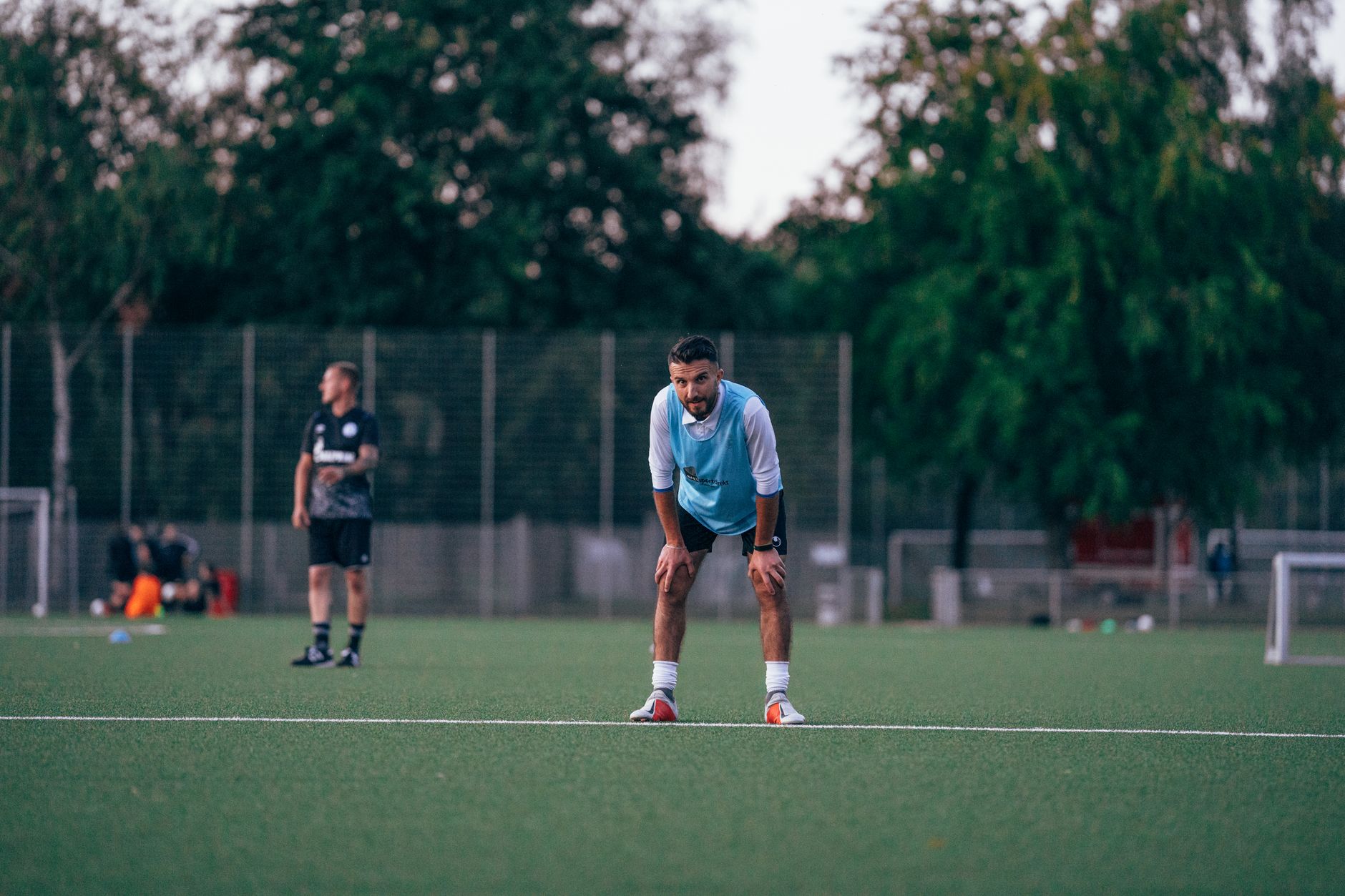 Un joueur en chasuble bleu clair, penché en avant les mains sur les genoux, sur un terrain d’entraînement de football en gazon synthétique.