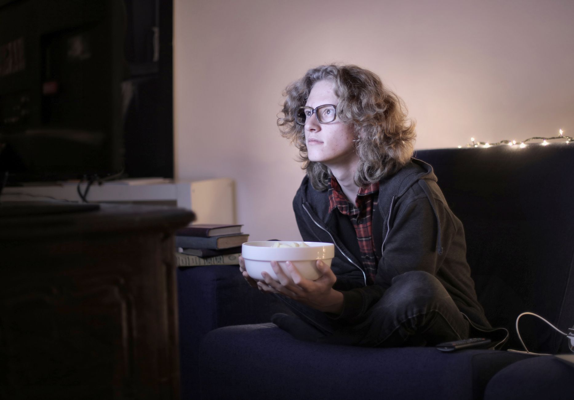 Une personne portant des lunettes regarde un écran de télévision depuis son canapé en tenant un bol.