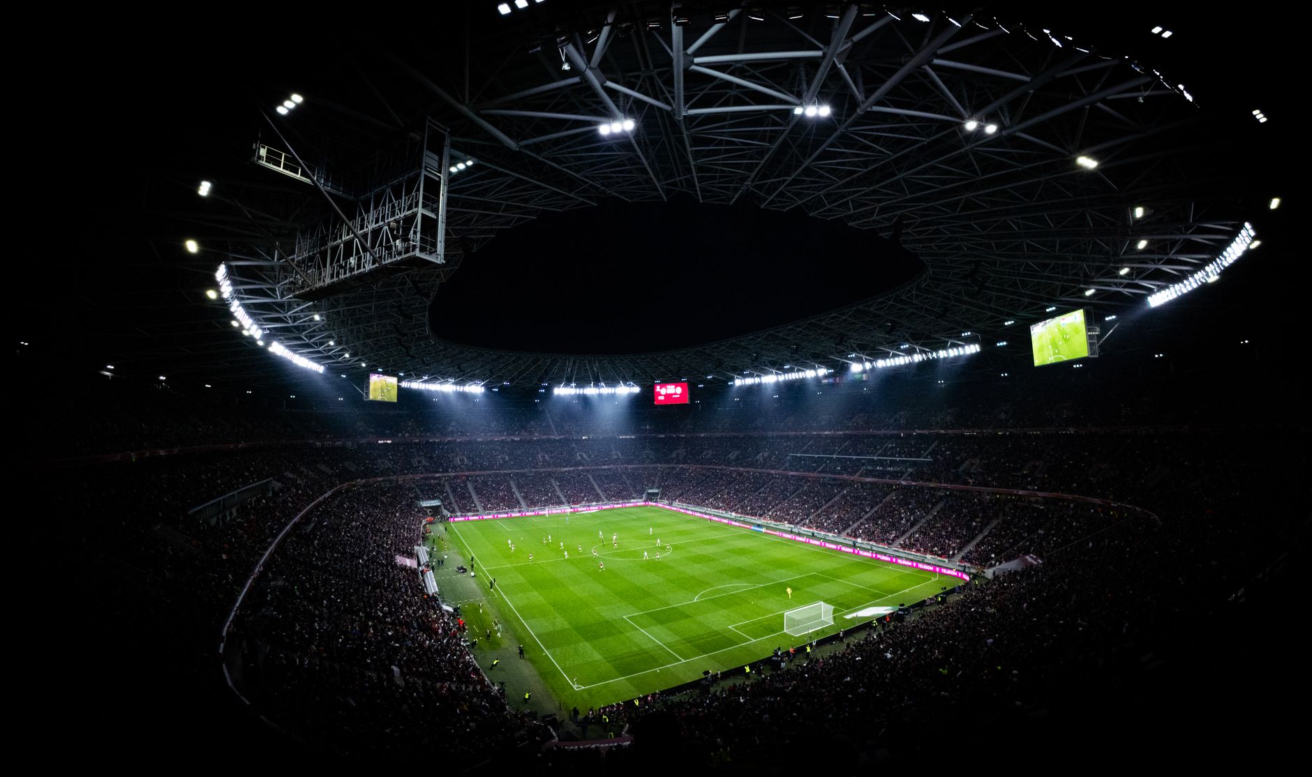 Vue en plongée d’un stade de football illuminé de nuit, montrant la pelouse verte et les tribunes pleines.